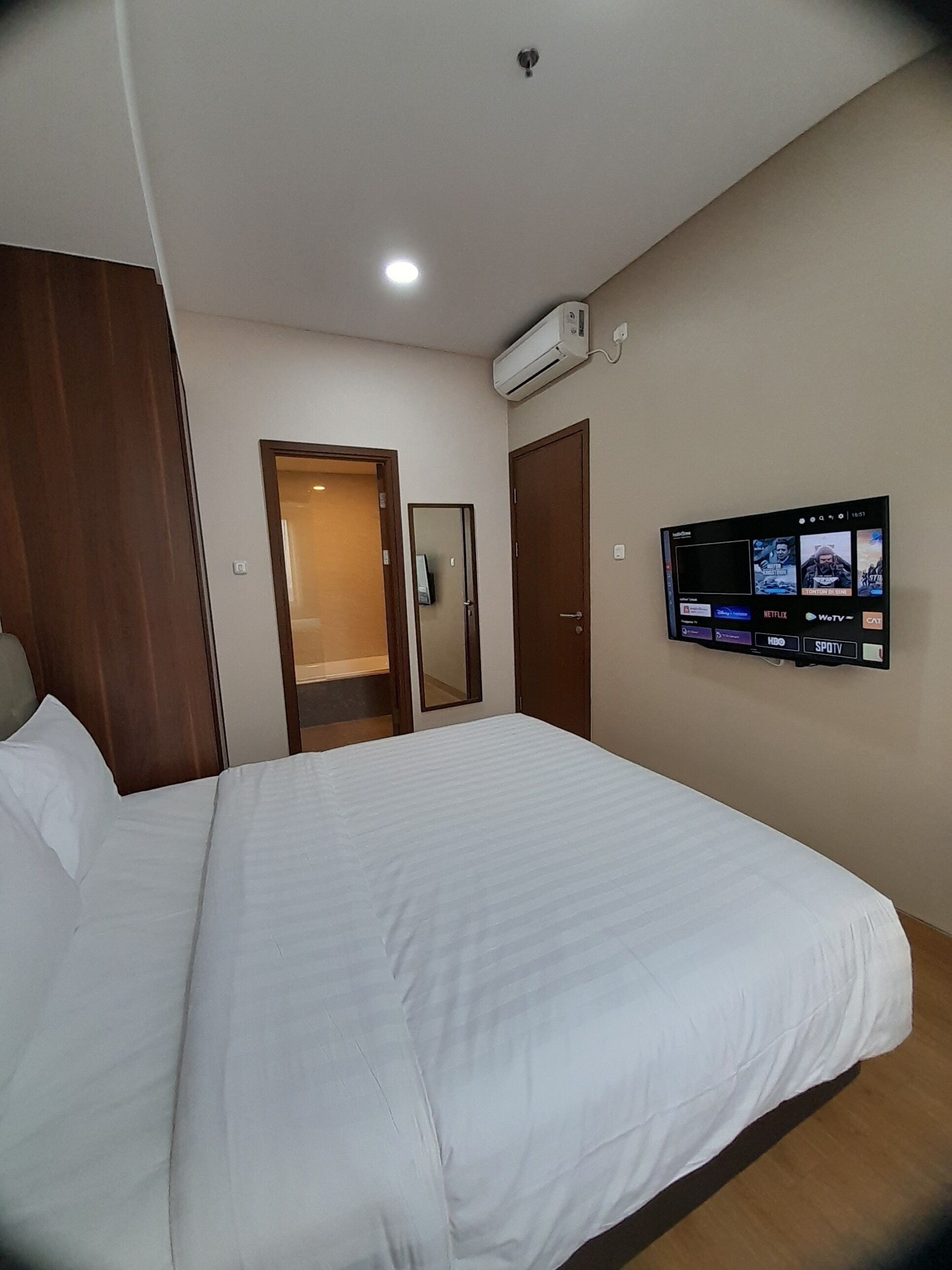 Habitación de hotel con una cama grande, un televisor de pantalla plana montado en la pared mostrando varios canales, un equipo de aire acondicionado sobre el televisor y un armario de madera.