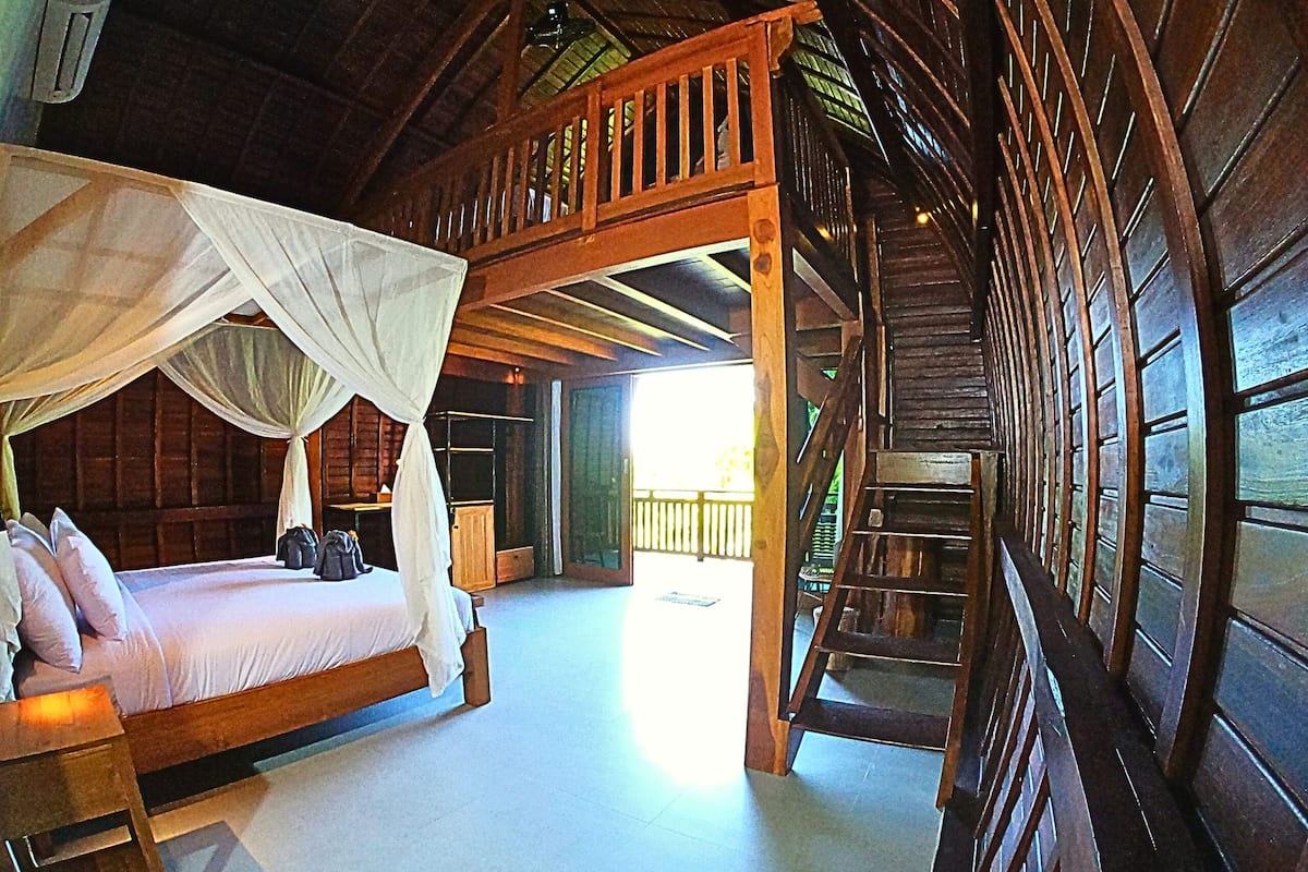 A wooden cabin with a bed under a canopy, a staircase, and a view of the outside.