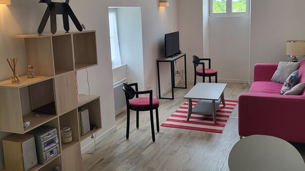 Un salon moderne avec un plancher en bois, un canapé rose, une table blanche et un téléviseur posé sur un meuble. On y trouve des étagères remplies de divers objets et une fenêtre donnant sur un paysage verdoyant.