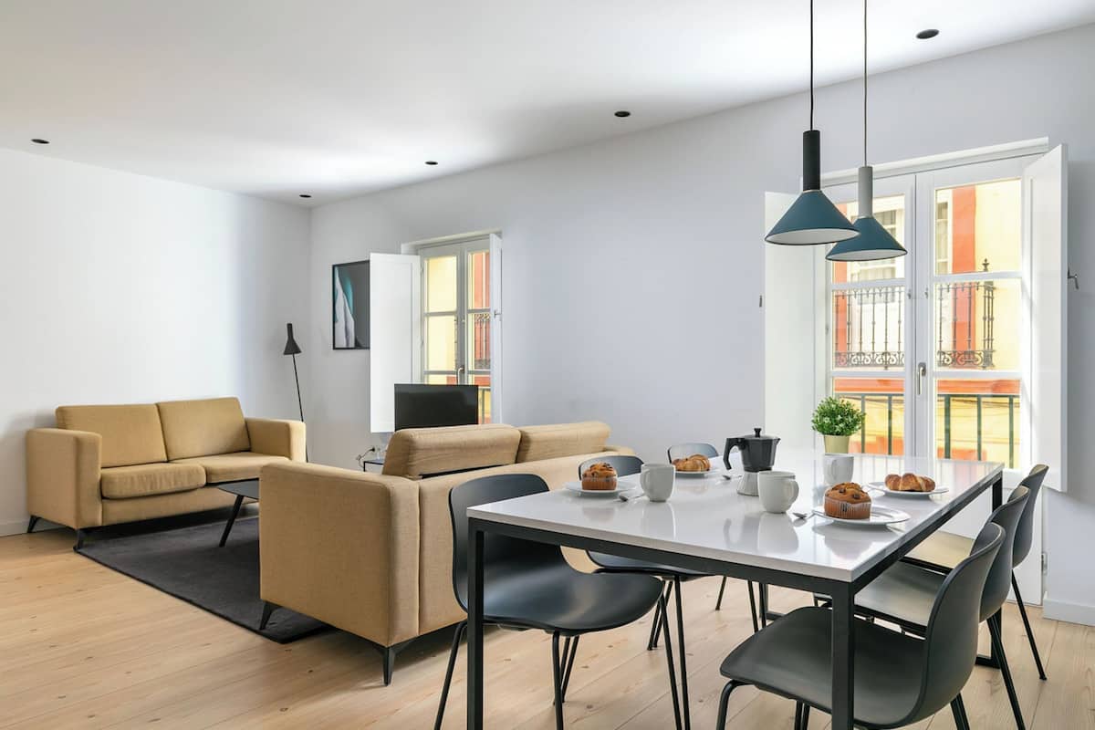 A modern living room with a sofa, dining table, and chairs. There is a window with a view of balconies.