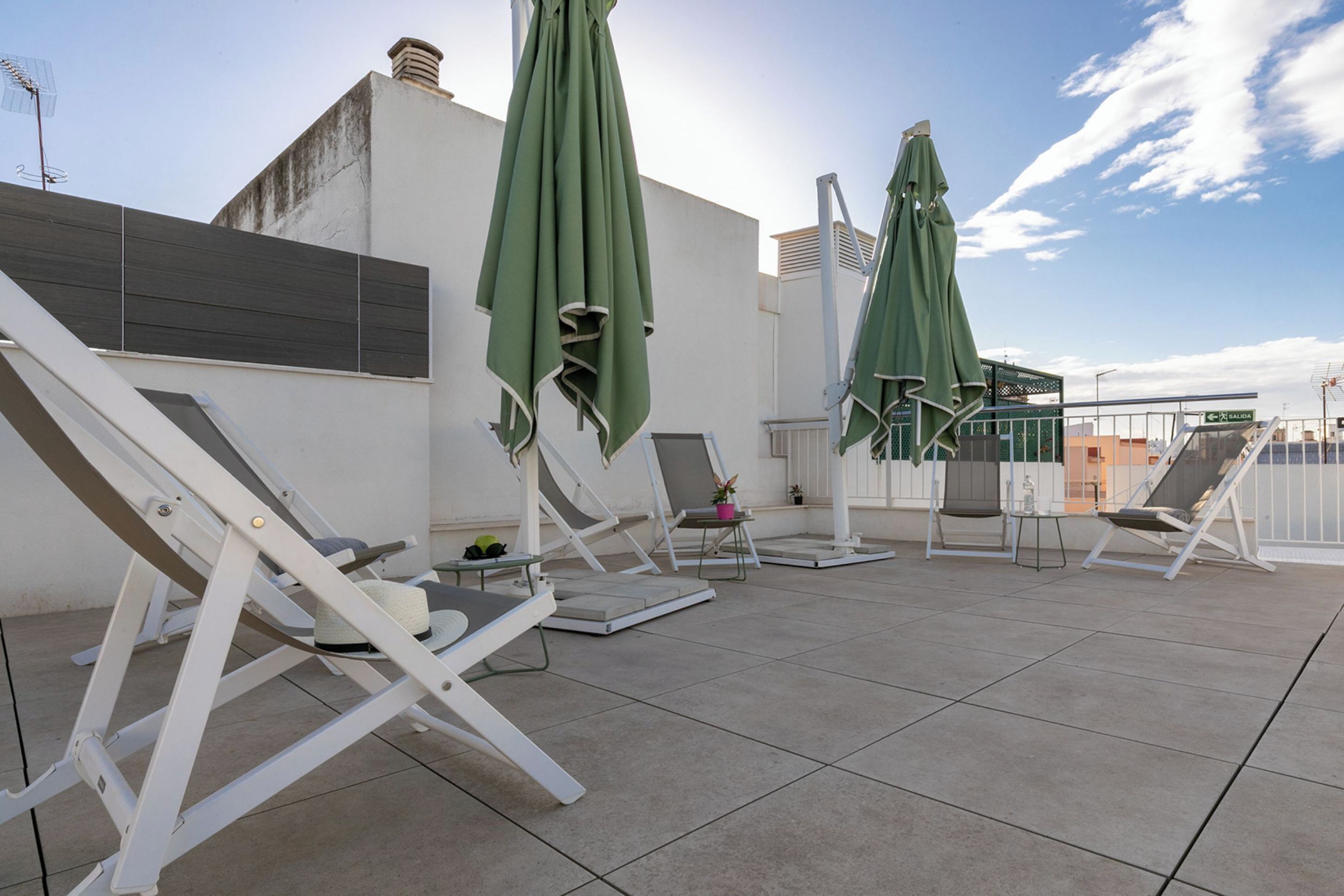 A rooftop terrace with multiple sun loungers and green umbrellas, a white building, and a clear sky.