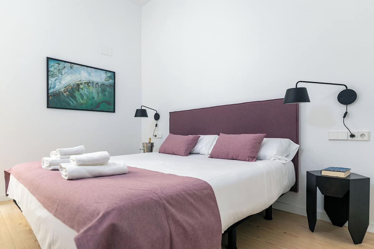 A modern bedroom with a bed, pillows, a bedside table, a lamp, and a painting on the wall.