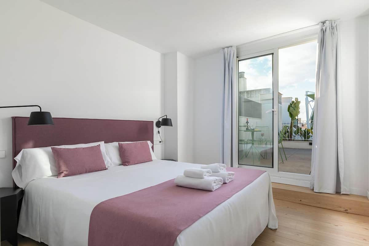 A modern hotel room with a large bed, a nightstand, a desk, and a balcony with a view of the city.