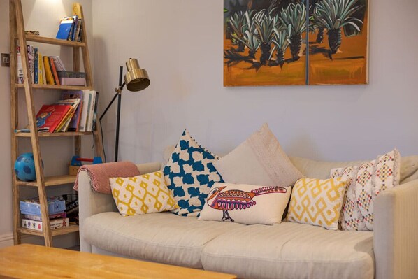 A living room with a beige sofa, colorful patterned cushions, a wooden bookshelf, and a floor lamp. - Trendy Hampstead Flat (RH7) (Greater London)
