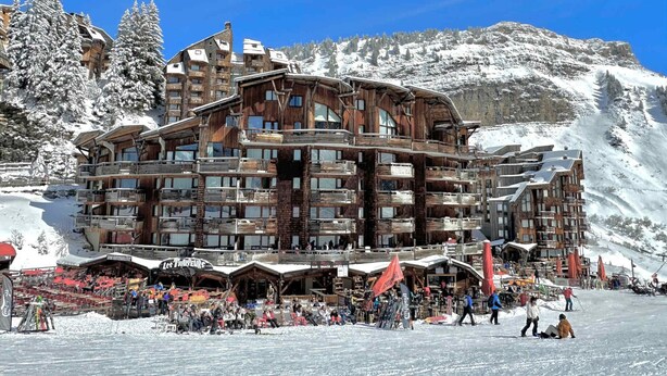 A multi-story wooden lodge with balconies, surrounded by snow-covered mountains and a ski slope.