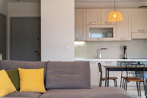 A modern living room with a grey sofa, yellow and green cushions, a dining area with a table and chairs, and a kitchen with light wooden cabinets and a hanging pendant light.