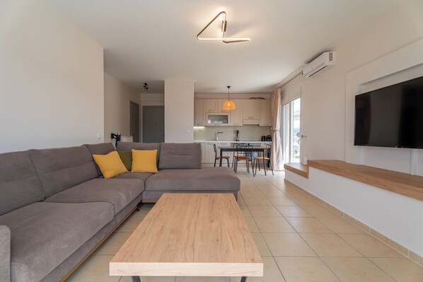 A modern living room with a sectional sofa, a wooden coffee table, a TV mounted on the wall, and a kitchen in the background.