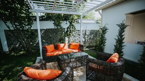 Ein möblierter Außenbereich mit Korbmöbeln und orangefarbenen Kissen, einer Pergola und einer Steinmauer.