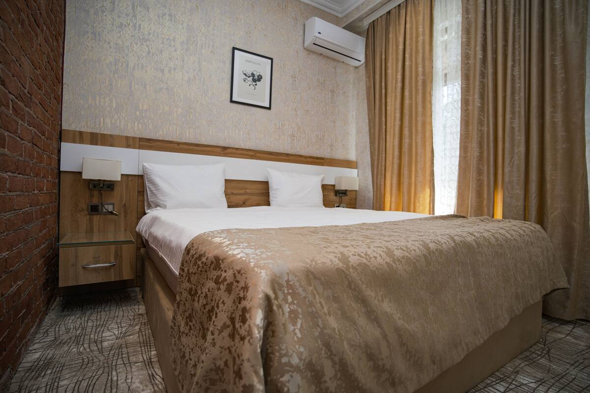 Quarto de hotel com uma cama grande, duas mesas de cabeceira, uma cabeceira de madeira, um ar condicionado fixado na parede e um quadro emoldurado.