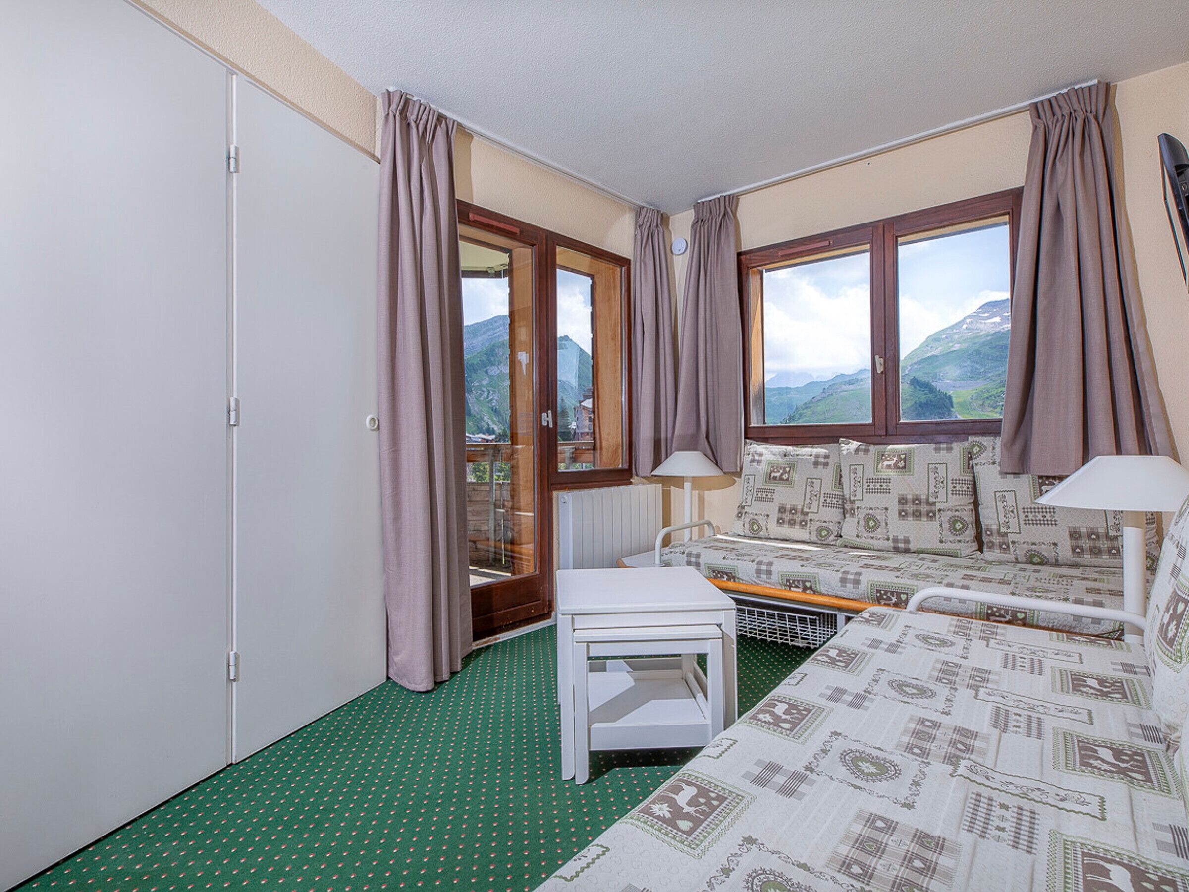 Une chambre avec un lit, une petite table et une vue sur les montagnes par les fenêtres.