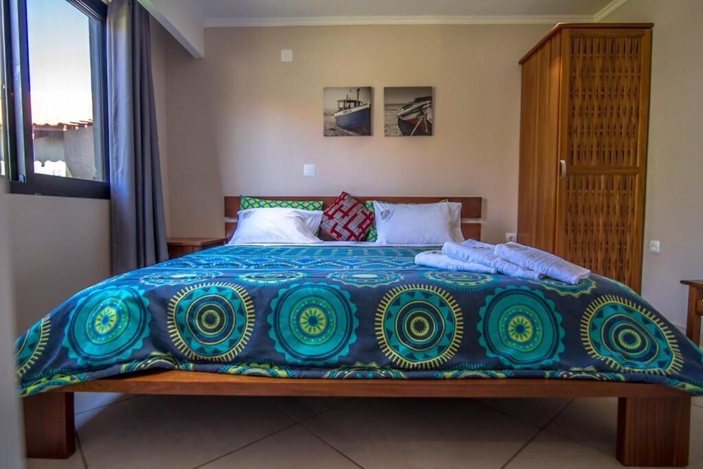 Un lit avec une couvre-lit à motifs, des oreillers et une armoire en bois, dans une pièce dotée d’une fenêtre et de deux tableaux accrochés au mur.