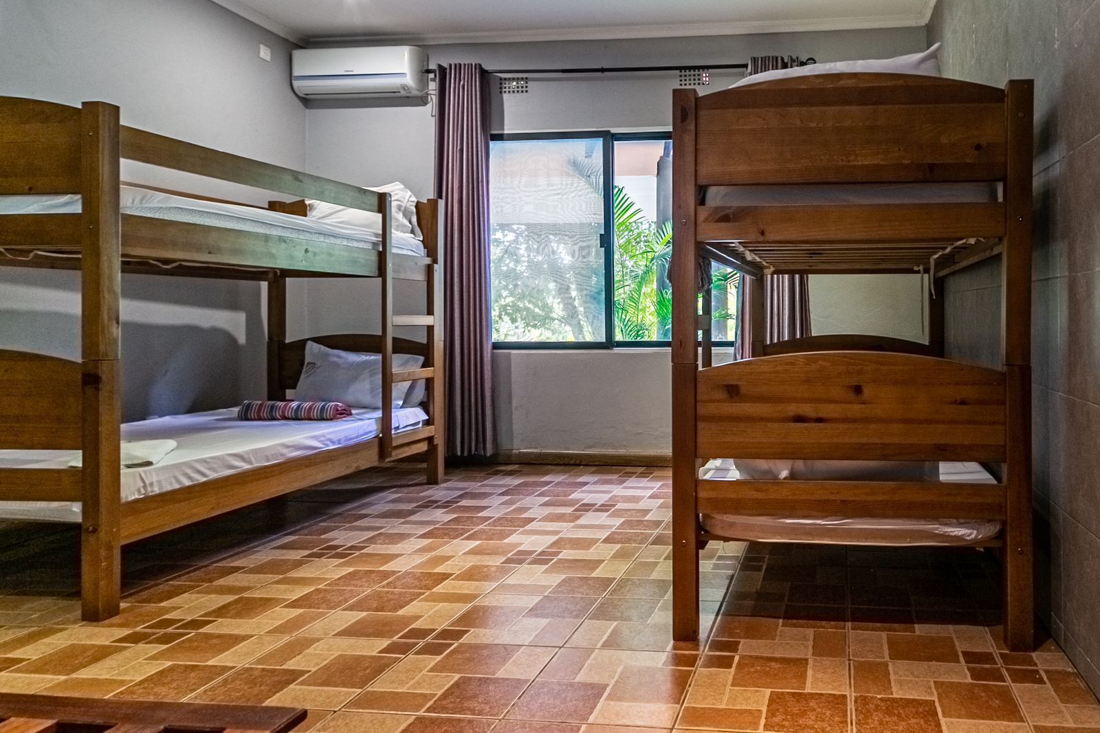 Una habitación con literas, suelo de baldosas, ventana con cortinas y un equipo de aire acondicionado.