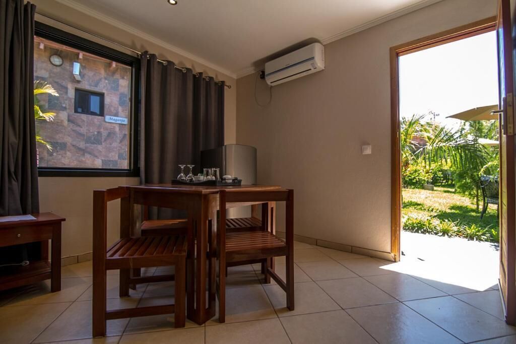 A room with a wooden dining table and chairs, a window with curtains, an air conditioning unit, and a view of a garden.