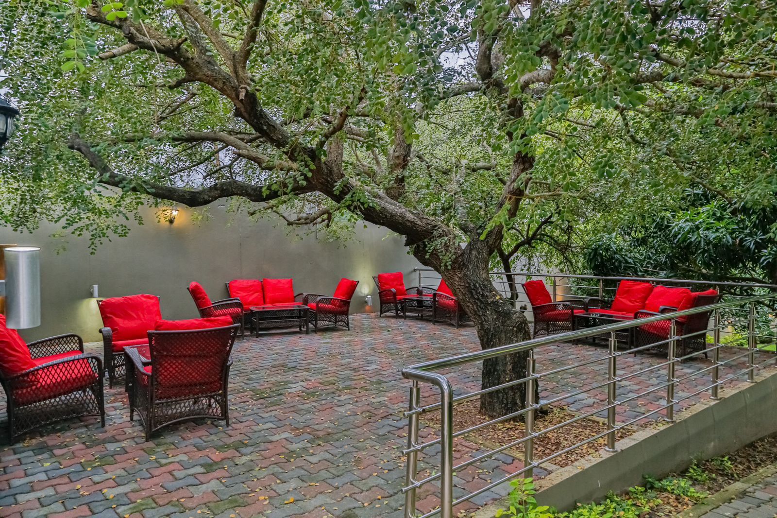 An outdoor seating area with wicker chairs and red cushions, surrounded by trees and a brick floor.