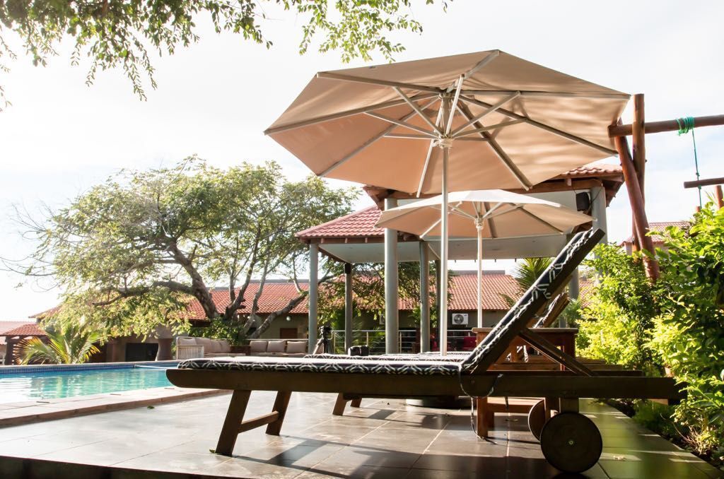Un espace aménagé au bord de la piscine, avec un fauteuil de relaxation et un parasol, entouré de verdure et bordé d’un bâtiment dont le toit est recouvert de tuiles.