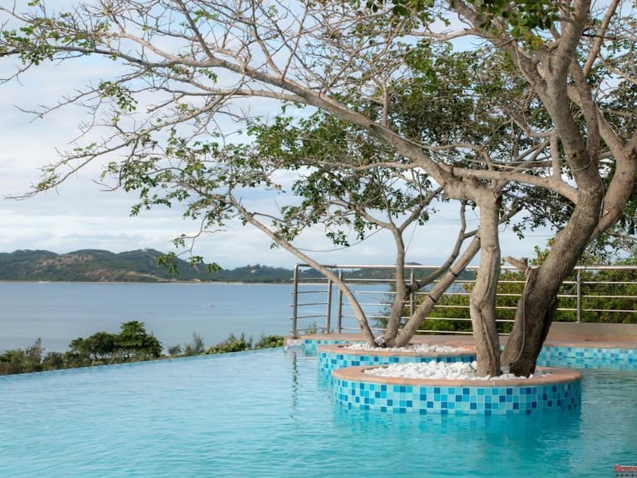 A pool with a tree in the center, surrounded by a tiled deck and a view of the sea.