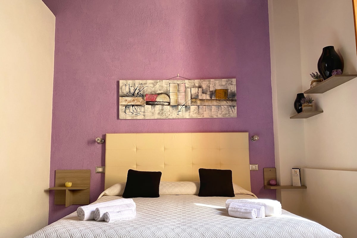 A hotel room with a large bed, a headboard, a wall art piece, and a shelf with decorative items.