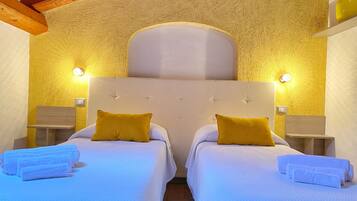 A double room with two beds, each with a white headboard and a single yellow pillow. There are two bedside tables with lamps, and towels are neatly folded on each bed.