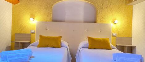 A double room with two beds, each with a white headboard and a single yellow pillow. There are two bedside tables with lamps, and towels are neatly folded on each bed.