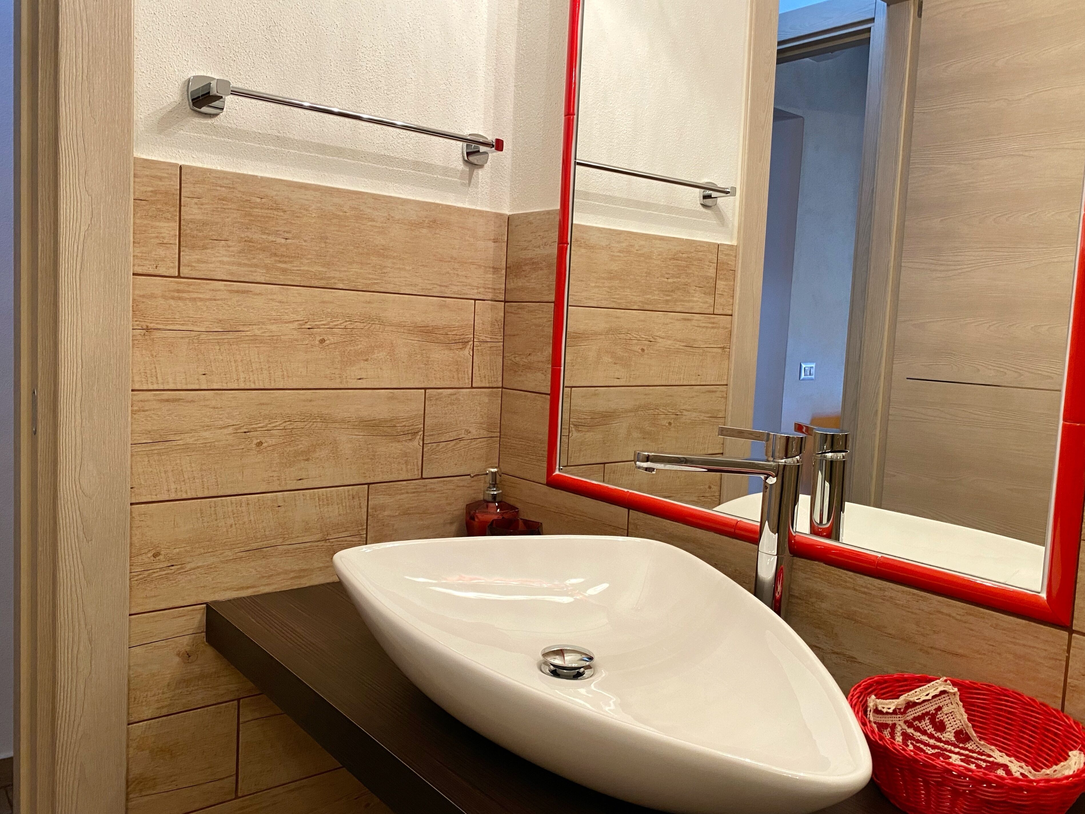A bathroom with a white sink, wooden panel walls, a red-framed mirror, and a red basket.