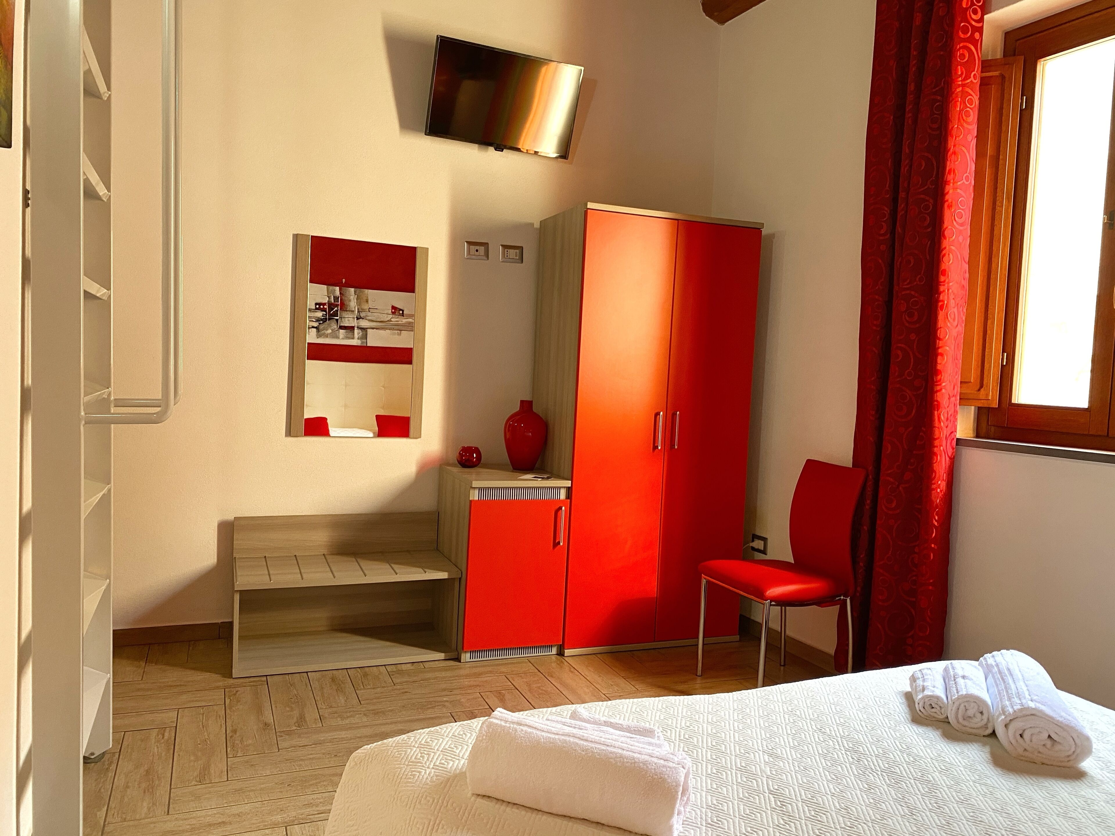 Habitación de hotel con cama, armario rojo, silla, mesita, espejo y televisor.