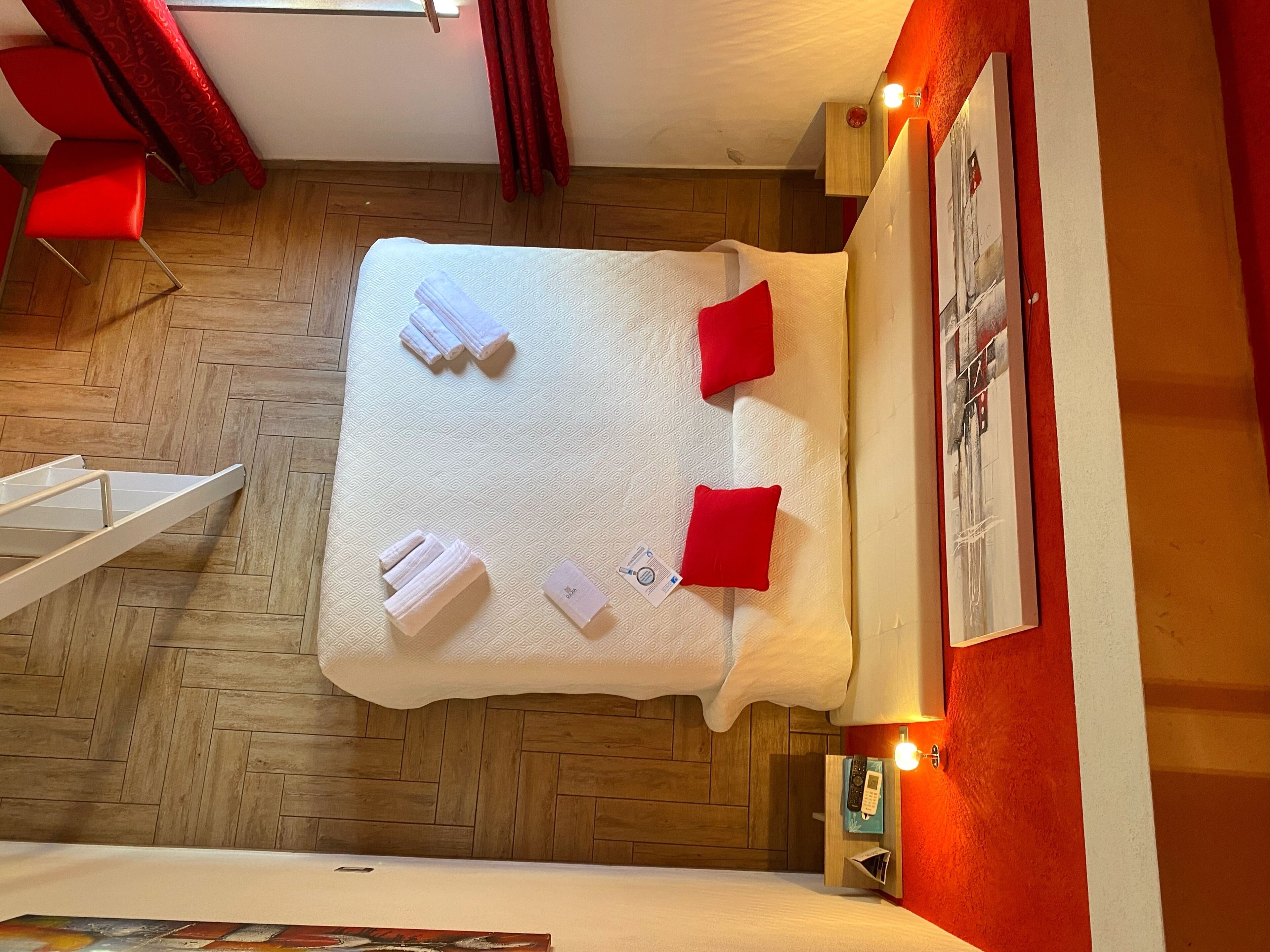 A neatly made bed with white linens and red pillows, a wooden floor, a red curtain, and a wall-mounted light fixture.
