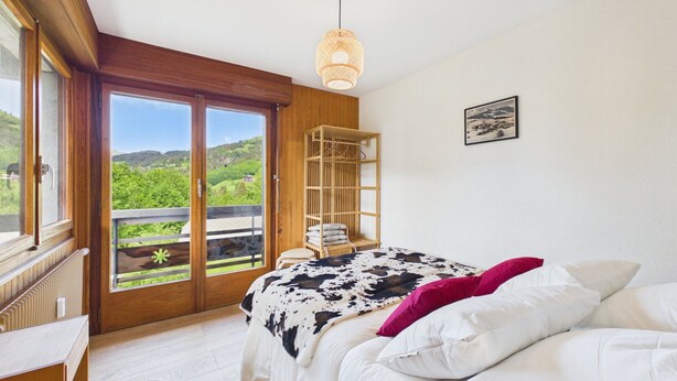 Een slaapkamer met een bed, een houten kledingkast, een raam met uitzicht op groen en een hanglamp.