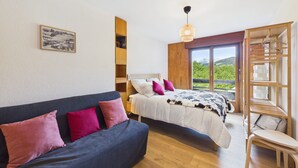 Een slaapkamer met een bed, een slaapbank, een houten plank, een hanglamp en een raam met uitzicht.