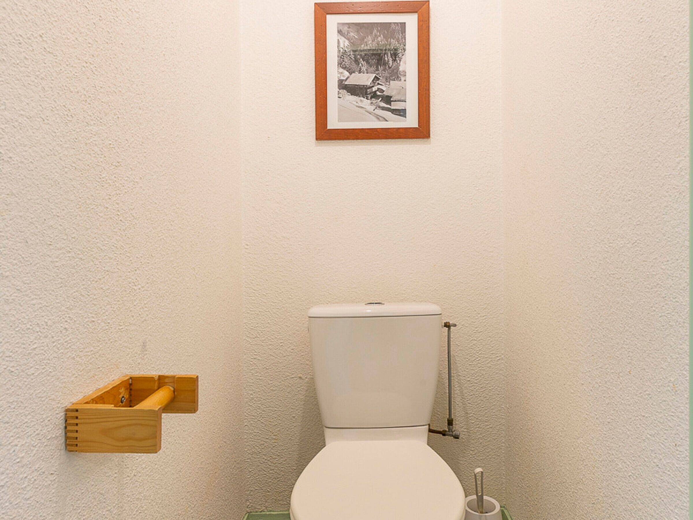 A bathroom with a toilet, a wooden shelf with a roll of toilet paper, and a framed picture on the wall.
