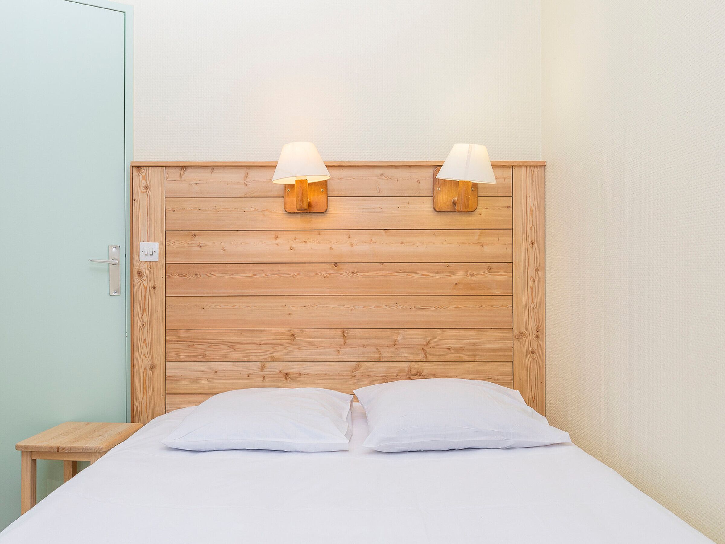 Een kamer met een houten hoofdbord, twee bedlampjes, een bed met wit beddengoed en een klein nachtkastje van hout.