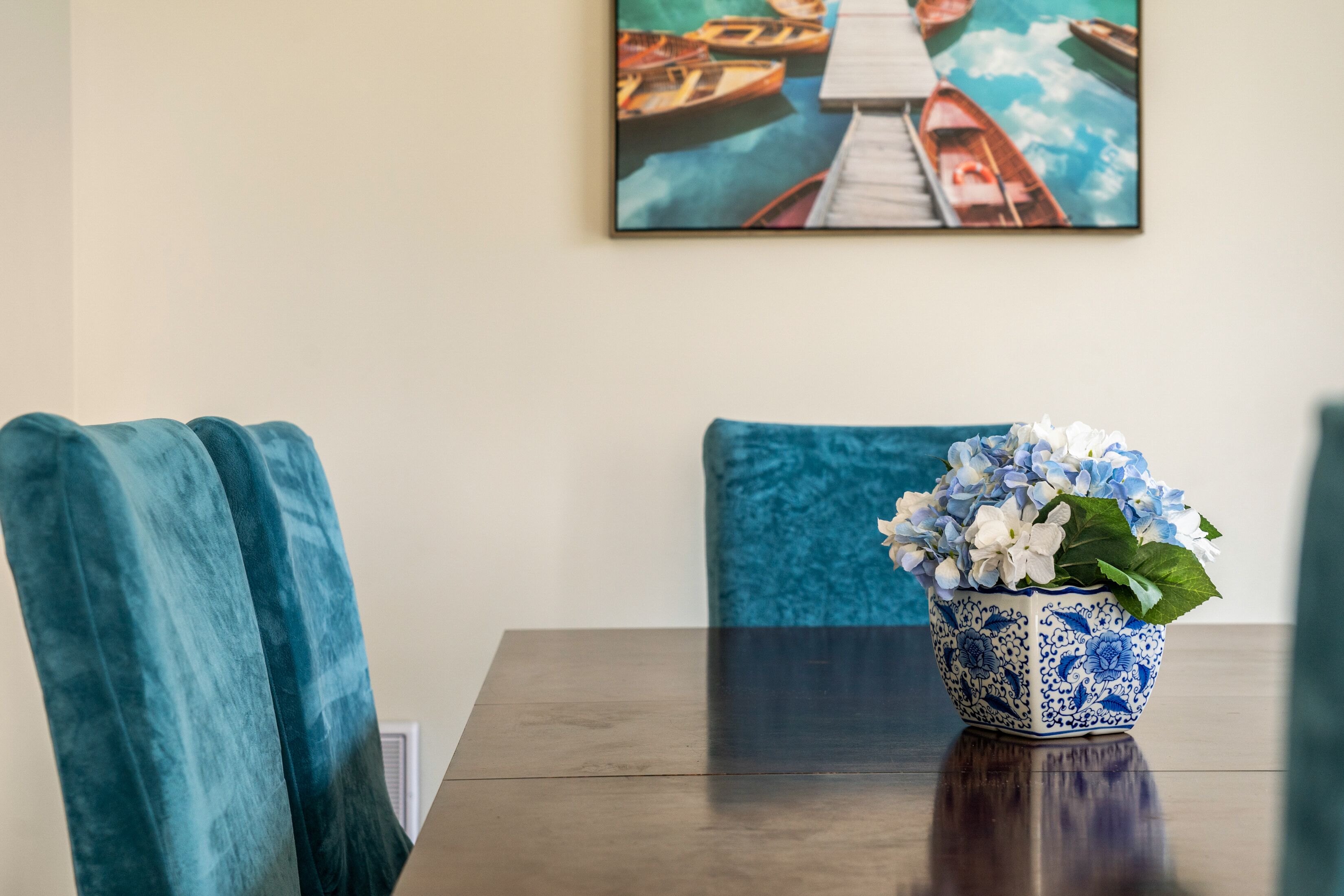 Un tavolo da pranzo con un vaso blu pieno di fiori, due sedie blu e un quadro con delle barche al muro.
