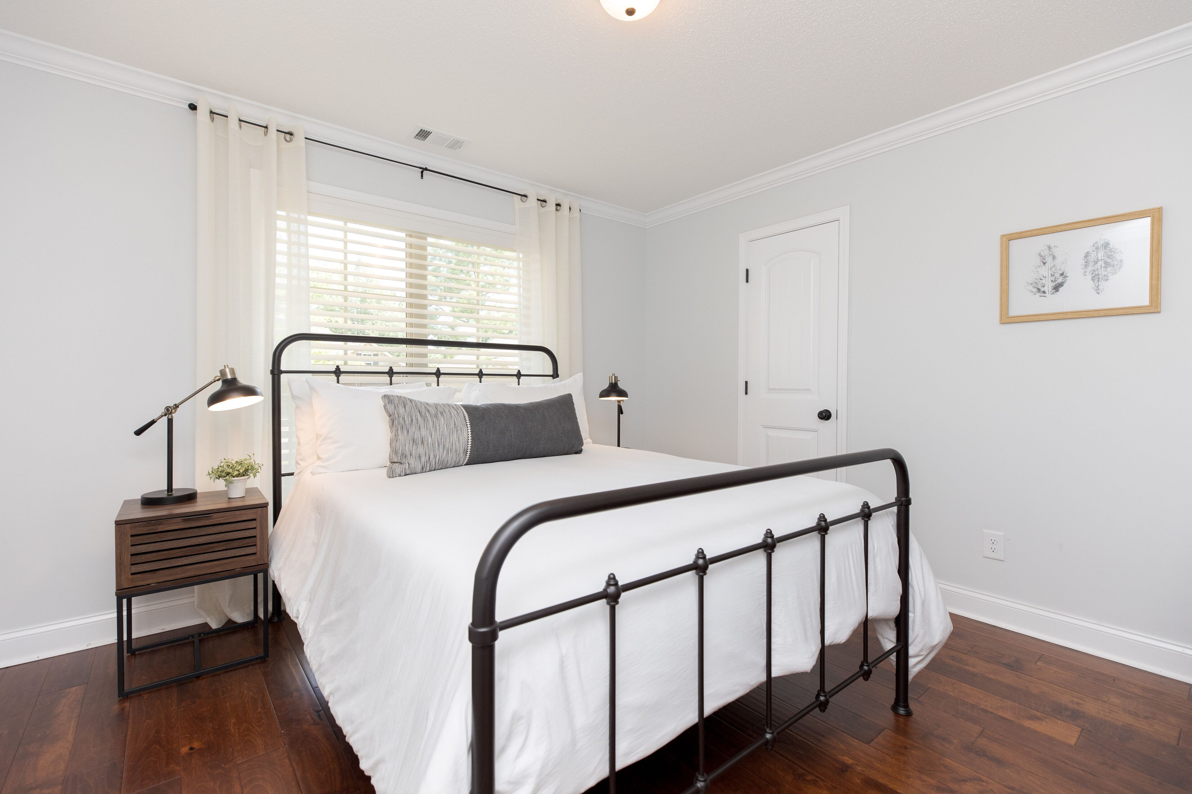 A bedroom with a metal-framed bed, white bedding, a bedside table with a lamp, and a window with curtains.