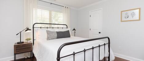 A bedroom with a metal-framed bed, white bedding, a bedside table with a lamp, and a window with curtains.
