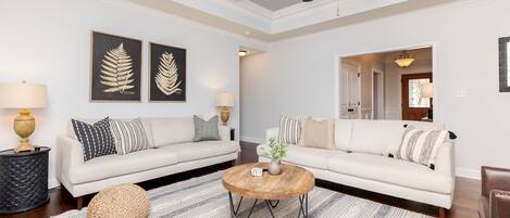 Un salón con un sofá blanco, una mesa de centro redonda y almohadones decorativos. Se observan obras de arte enmarcadas en la pared y un ventilador de techo.