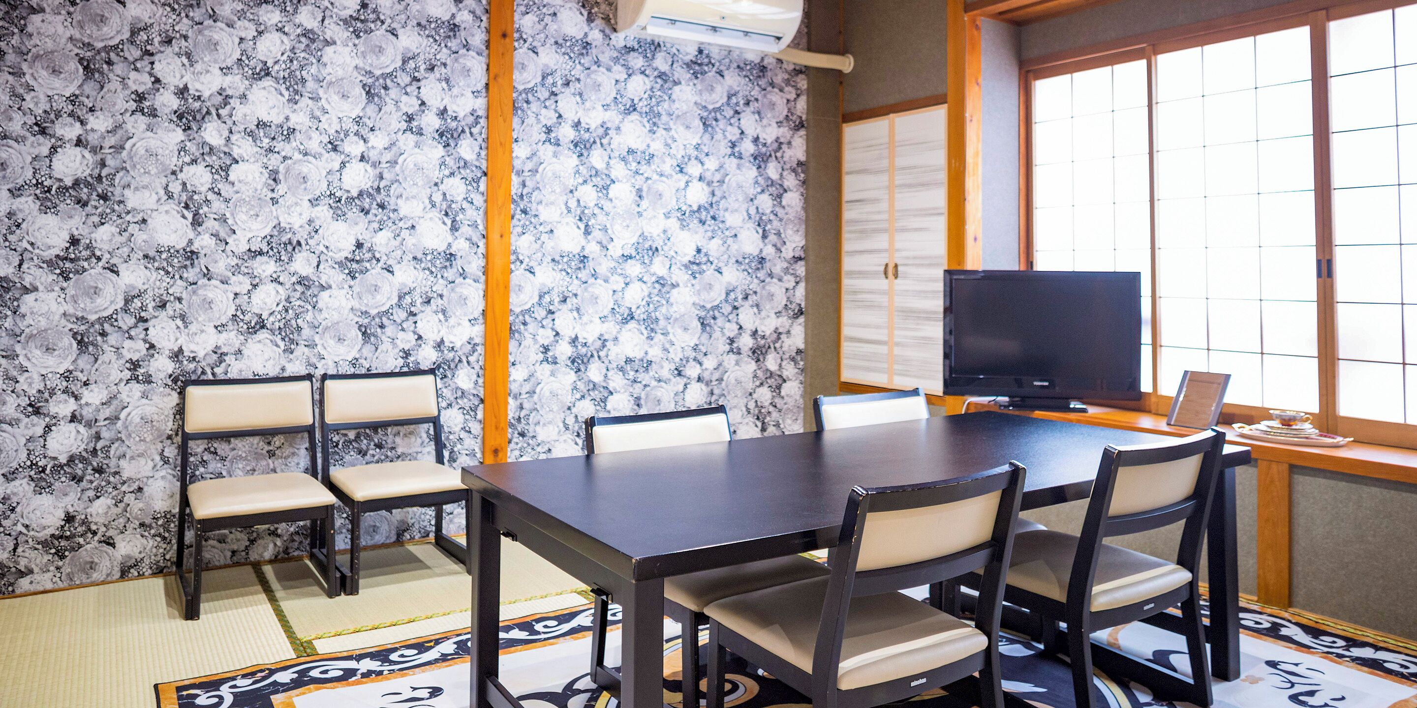 Un salón de conferencias con una mesa de madera oscura, sillas color beige, un televisor de pantalla plana y piso de esteras de tatami.