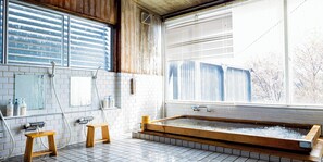Un baño con una bañera de madera, paredes de azulejos blancos, una ventana con persianas y un taburete pequeño de madera.