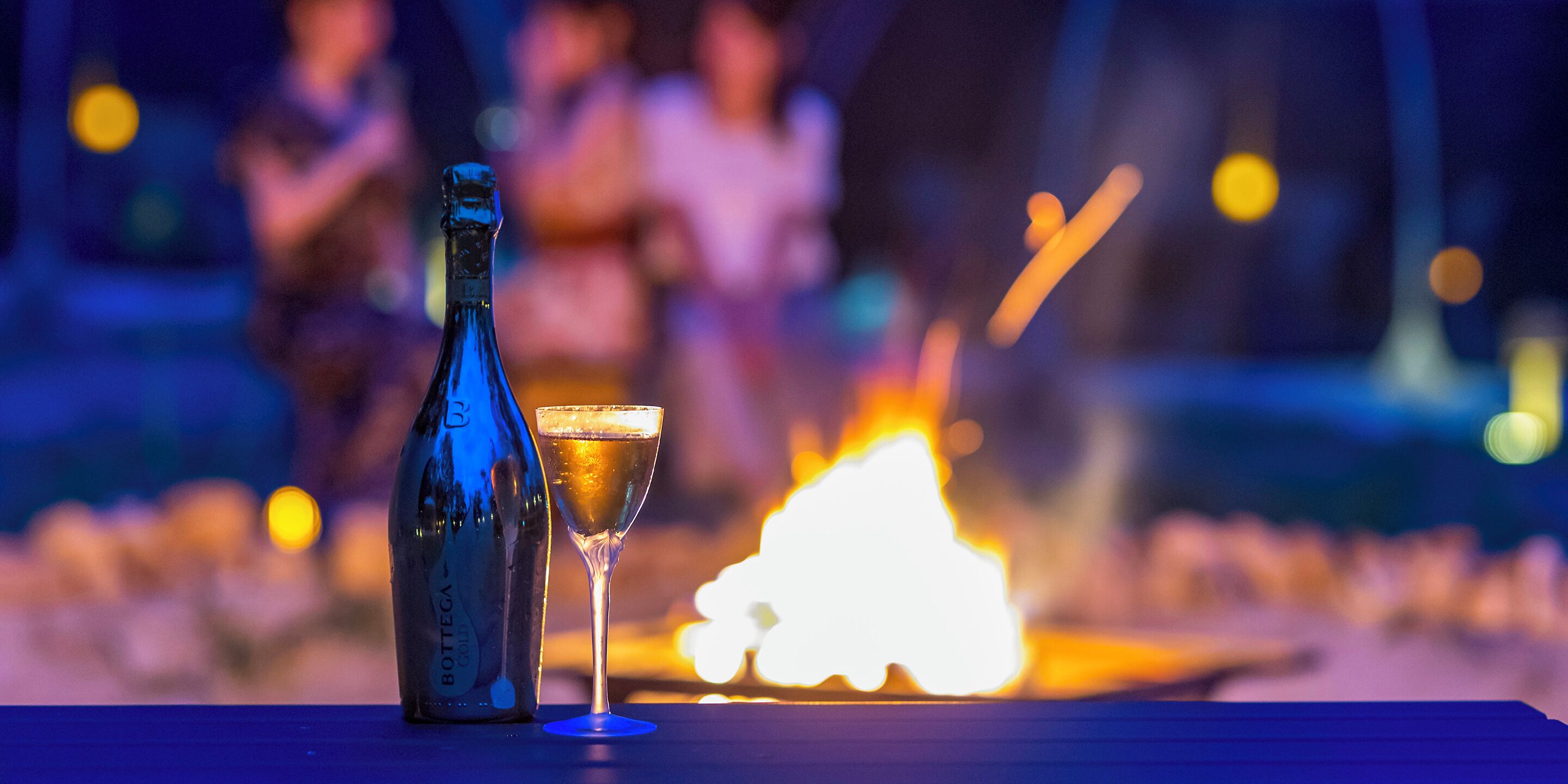 A bottle of Bottega wine and a glass of wine on a table with a fire in the background.