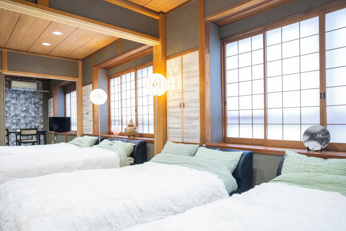 Una habitación tradicional japonesa con suelo de tatami, techo de madera bajo y puertas correderas shoji. Hay dos camas con ropa blanca y una mesita con un televisor.