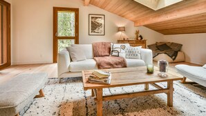A cozy loft bedroom with a wooden ceiling, a large window, a sofa, a coffee table, and a side table with a lamp.