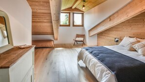 A bedroom with a sloped ceiling, wooden beams, a bed, a chair, and a window.