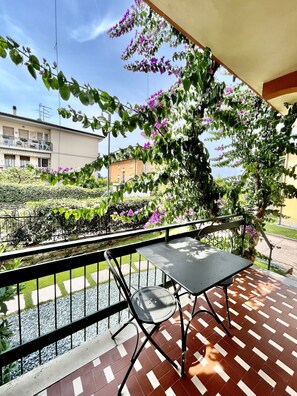 A balcony with a table and chair, overlooking a garden with purple flowers and a building in the background. - Bougainvillea Residence (Torri del Benaco)