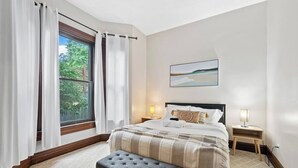 Une chambre à coucher avec un lit, deux tables de chevet avec des lampes, un banc et une fenêtre avec des rideaux.