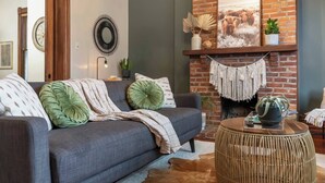 Un salon chaleureux avec un canapé gris, des coussins verts, une table basse ronde en osier et une cheminée en briques surmontée d’un tableau encadré.