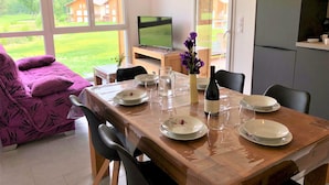 A dining area with a wooden table set for four, a TV in the background, and a view of a green landscape through large windows. - Modern apartment with terrace and private parking (Barcelonnette)