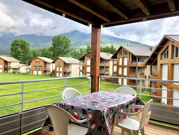A covered outdoor area with a table and chairs, overlooking a row of wooden buildings with balconies, set against a mountainous backdrop. - Modern apartment with terrace and private parking (Barcelonnette)