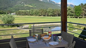A table set with plates, cutlery, a bottle, and a mug, overlooking a scenic landscape with mountains and a golf course. - Modern apartment with terrace and private parking (Barcelonnette)