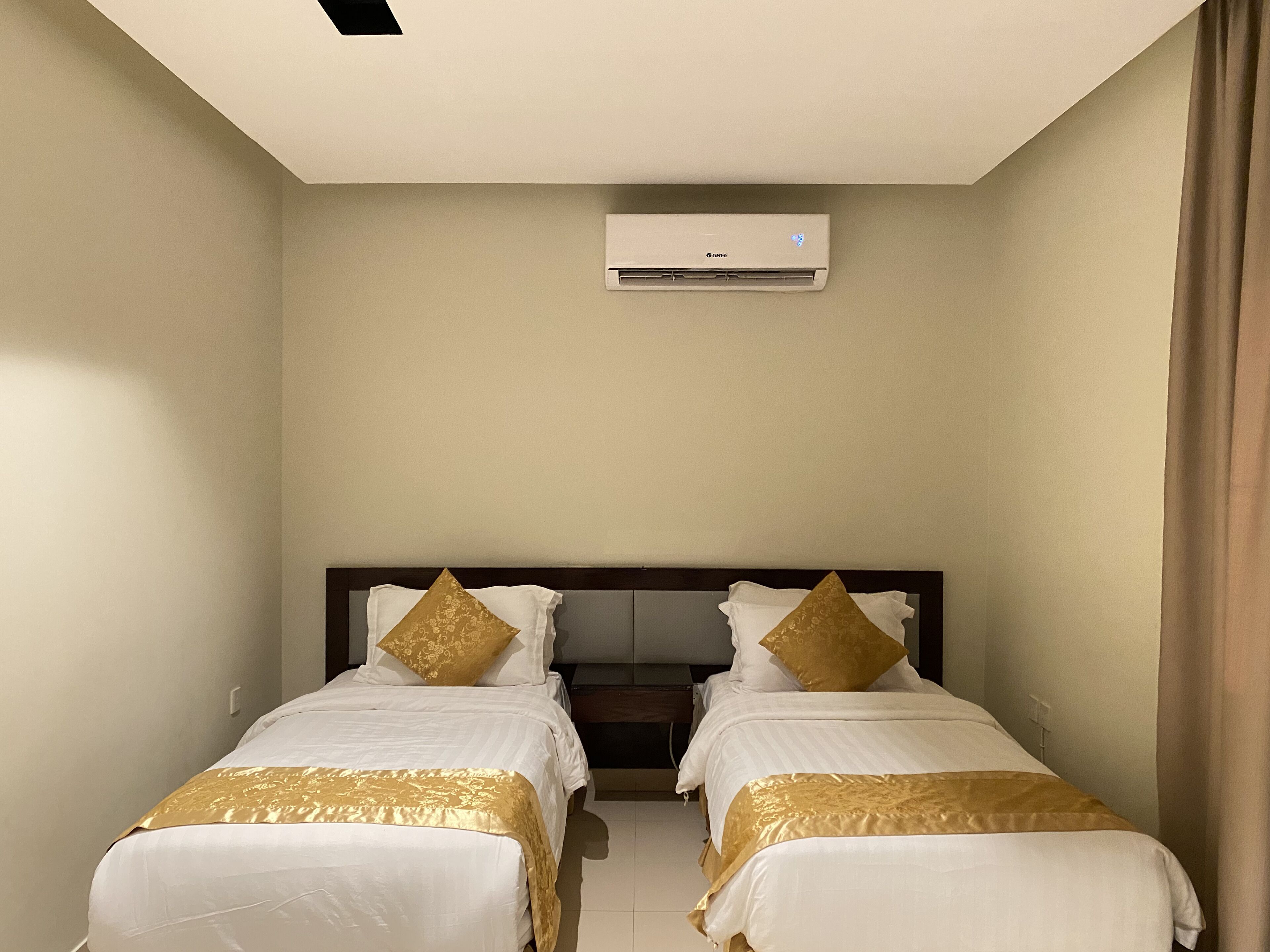 A hotel room with two single beds, a dark headboard, an air conditioning unit, and a curtain on the right side.
