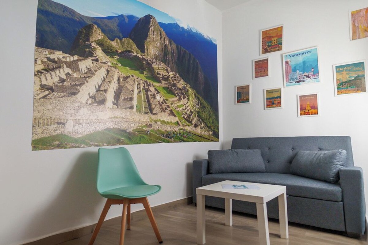 Una habitación con un cartel grande de Machu Picchu, un sofá gris, una silla color turquesa y una mesita.