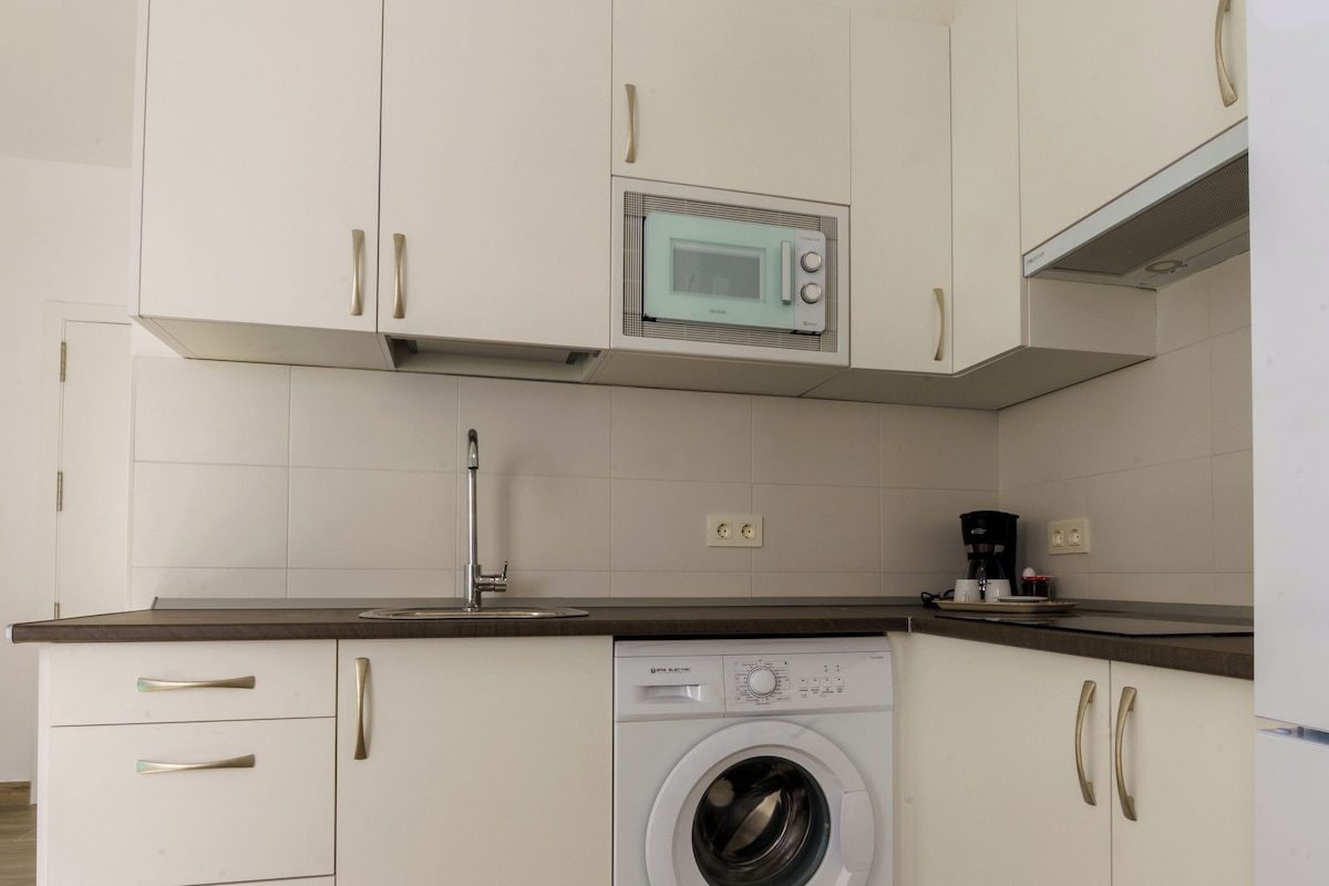 Cocina compacta con armarios blancos, lavadora, microondas y cafetera.