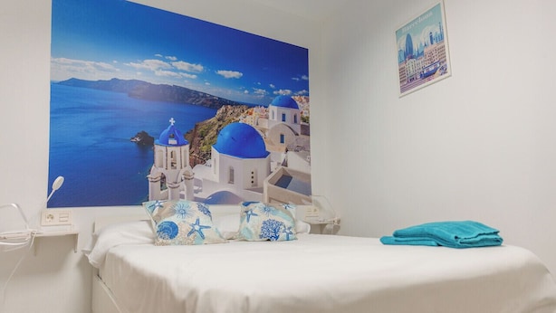 A hotel room with a large wall mural of a coastal scene, a neatly made bed with blue and white pillows, and a folded blue towel on the bed. - APARTAMENTOS MAPAMUNDI (Badajoz)